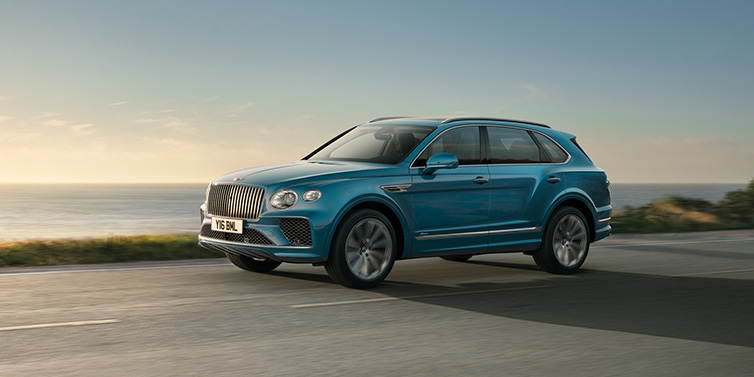 Bentley Warszawa Bentley Bentayga Azure SUV in Topaz blue paint driving dynamically by the ocean with 22 inch 10 spoke directional wheels