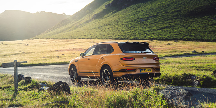 Bentley Warszawa Bentley Bentayga S SUV rear three quarter in Sunburst Gold paint on a road surrounded by green hills