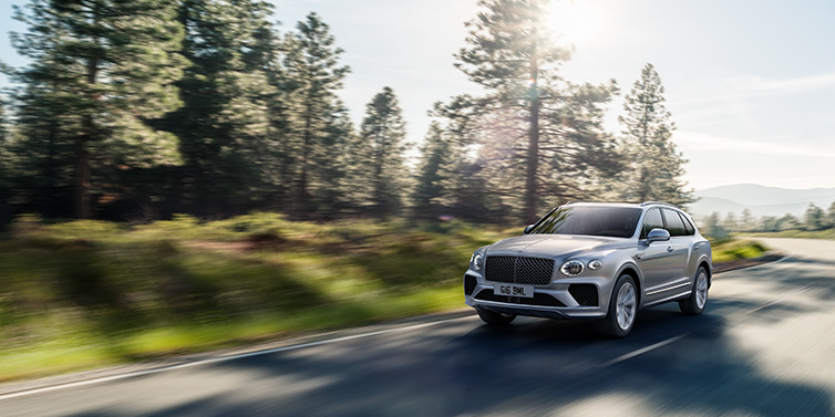 Bentley Warszawa Bentley Bentayga SUV front three quarter in Moonbeam silver paint driving dynamically along a tree-lined road